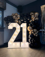 A large illuminated marquee number 21 stands on the floor in front of a balloon arch made of black, gray, and metallic gold balloons, in a decorated indoor room.