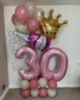 A 30th birthday balloon decoration set featuring two large pink foil '30' balloons, a pink champagne bottle balloon, a gold crown balloon, and several pink, white, and silver balloons arranged against a plain indoor wall.