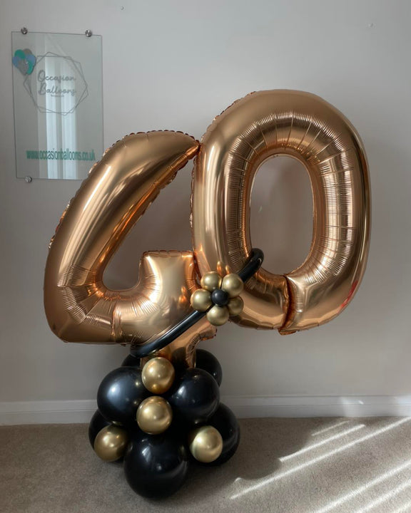 A large metallic rose gold foil balloon shaped like the number 40 stands on a balloon cluster base with black and gold balloons, set against a plain indoor wall.