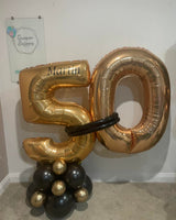 Gold and black foil balloon numbers spelling '50' with a black ring prop, displayed on a carpeted floor against a white wall with a branded sign in the background.