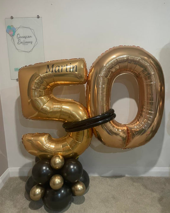 Gold and black foil balloon numbers spelling '50' with a black ring prop, displayed on a carpeted floor against a white wall with a branded sign in the background.