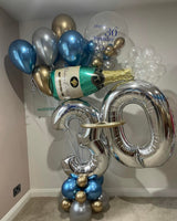 A birthday balloon bouquet arrangement in a room corner, featuring metallic silver number 30 balloons, blue and silver balloons, gold and silver accent balloons, a clear balloon printed with 'Happy 30th Birthday', and a champagne bottle decoration.