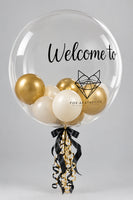 A large clear balloon printed with the words 'Welcome to' and a black geometric fox logo, filled with smaller metallic gold, cream, and white balloons. The balloon is tied with a black and gold ribbon at the bottom against a plain light background.