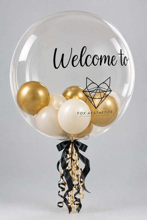 A large clear balloon printed with the words 'Welcome to' and a black geometric fox logo, filled with smaller metallic gold, cream, and white balloons. The balloon is tied with a black and gold ribbon at the bottom against a plain light background.