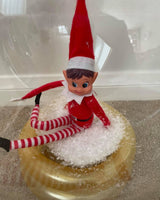 Elf on the Shelf sitting on a gold lid with snow-like material.