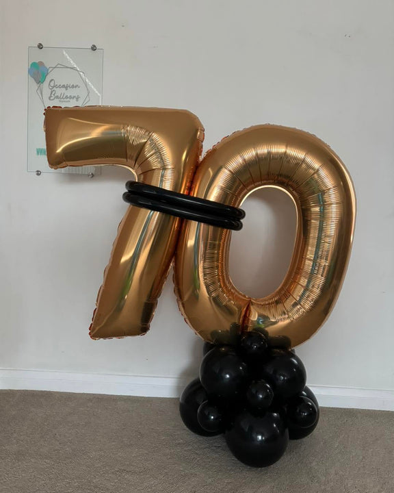 Gold 70 foil number balloons joined with black balloon bands on a freestanding black balloon base.