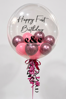 A large clear round balloon with 'Happy First Birthday e&e boutique' printed on it, filled with pink and metallic rose-gold balloons, tied with a pink and black ribbon and curling streamers on a white background.