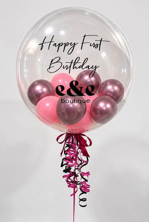 A large clear round balloon with 'Happy First Birthday e&e boutique' printed on it, filled with pink and metallic rose-gold balloons, tied with a pink and black ribbon and curling streamers on a white background.