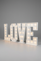 Large illuminated marquee letters spelling LOVE displayed on the floor in a decorated indoor room with white brick walls, wooden flooring, balloons, and a blue curtain.