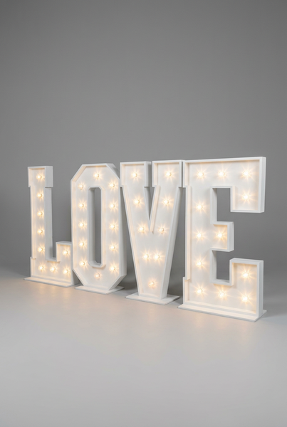 Large illuminated marquee letters spelling LOVE displayed on the floor in a decorated indoor room with white brick walls, wooden flooring, balloons, and a blue curtain.