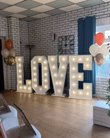 Freestanding white light-up LOVE letters with warm lights, flanked by orange, white and metallic helium balloon bouquets.