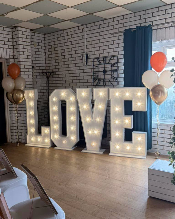 Freestanding white light-up LOVE letters with warm lights, flanked by orange, white and metallic helium balloon bouquets.