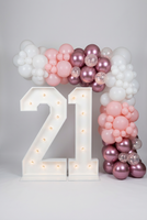 Large illuminated marquee numbers '21' on a carpeted floor in a room, styled with a balloon garland arch in pink, rose gold, and white.