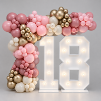 A decorative party setup featuring a balloon garland arch in shades of pink, blush, champagne gold, and white, alongside large illuminated white marquee numbers showing 18, on a light gray studio background.