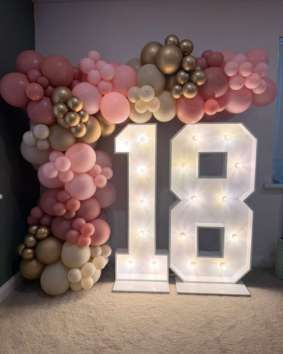 A party decoration setup featuring large illuminated marquee numbers '18' on a white carpet, framed by a balloon garland in blush pink, champagne gold, and cream tones against a dark gray wall.