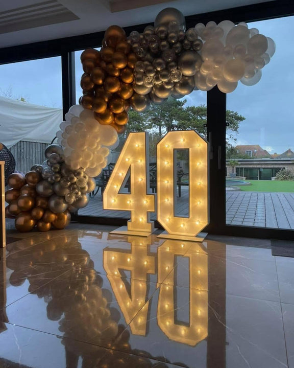 Illuminated 40 numbers beneath a sweeping gold, silver, white and confetti balloon garland at an indoor birthday venue.