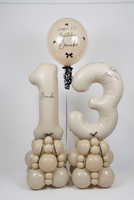 A custom birthday balloon arrangement for a 13th birthday featuring a large number 13 balloon, a round balloon reading 'Happy 13th Birthday Brooke', and two balloon towers shaped like animals, all in a warm cream color with black lettering and bows, set against a plain white wall.