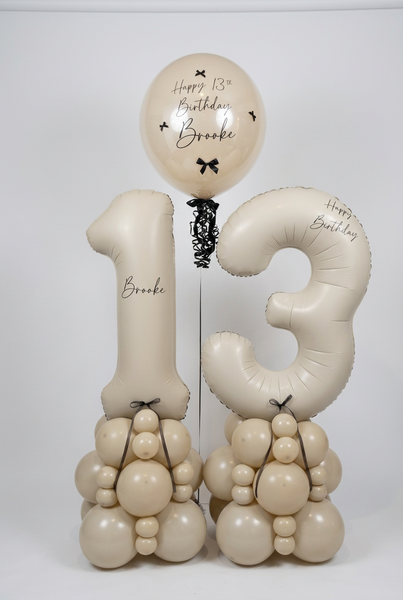 A custom birthday balloon arrangement for a 13th birthday featuring a large number 13 balloon, a round balloon reading 'Happy 13th Birthday Brooke', and two balloon towers shaped like animals, all in a warm cream color with black lettering and bows, set against a plain white wall.