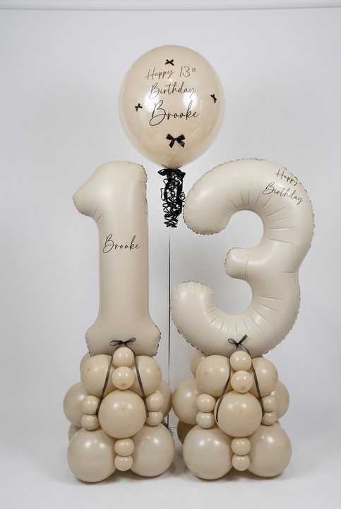 A custom birthday balloon arrangement for a 13th birthday featuring a large number 13 balloon, a round balloon reading 'Happy 13th Birthday Brooke', and two balloon towers shaped like animals, all in a warm cream color with black lettering and bows, set against a plain white wall.