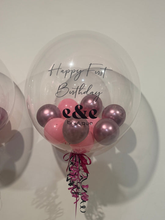 Clear company logo bubble balloon with pink and metallic berry mini balloons, e&e boutique branding and Happy First Birthday wording.