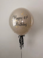 A clear round birthday balloon with 'Happy 41st Birthday' printed in black script and scattered gold foil confetti, tied with a black ribbon at the bottom, against a plain light gray background.