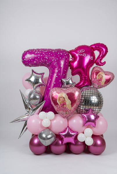 A pink and purple birthday balloon bouquet featuring a large number 7 balloon, a unicorn-shaped balloon with the name 'Luna', a Barbie heart balloon, silver disco ball balloons, and pink and white latex balloons, arranged against a plain wall.