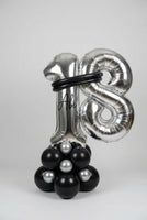 A large metallic silver foil balloon decoration shaped like the number 18, with a black circular band around the upper area and a cluster of black and silver balloons at the base, photographed indoors against a white wall.