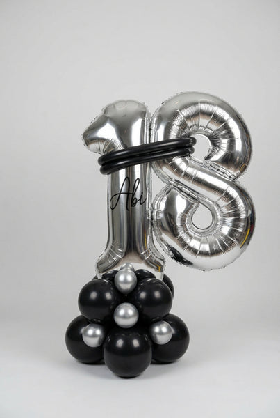 A large metallic silver foil balloon decoration shaped like the number 18, with a black circular band around the upper area and a cluster of black and silver balloons at the base, photographed indoors against a white wall.