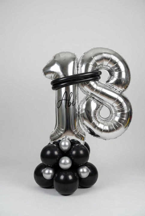 A large metallic silver foil balloon decoration shaped like the number 18, with a black circular band around the upper area and a cluster of black and silver balloons at the base, photographed indoors against a white wall.