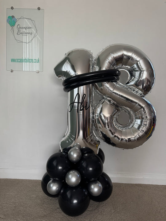 A large metallic silver foil balloon decoration shaped like the number 18, with a black circular band around the upper area and a cluster of black and silver balloons at the base, photographed indoors against a white wall.