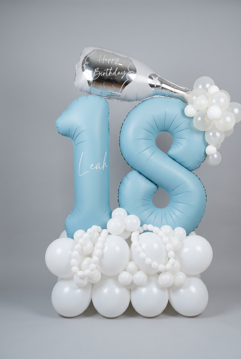 Blue 18 foil balloons personalised with Leah, topped with a silver birthday bottle balloon and clear bubbles, on a blue and white balloon base.