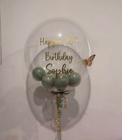 A large clear round balloon with gold foil text reading 'Happy 36th Birthday Sophie' and a small butterfly decoration, filled with pale green balloons at the bottom, tied against a plain light wall.