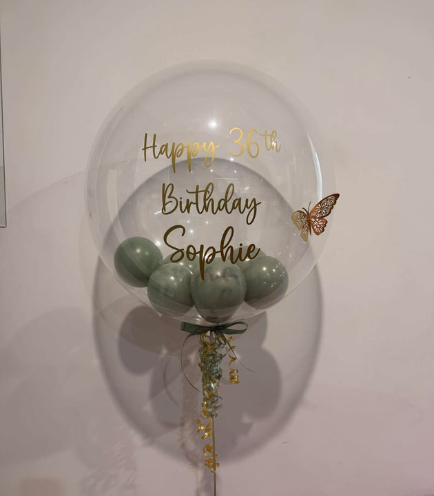 A large clear round balloon with gold foil text reading 'Happy 36th Birthday Sophie' and a small butterfly decoration, filled with pale green balloons at the bottom, tied against a plain light wall.