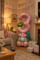 A large metallic pink number 3 birthday balloon decoration with dinosaur foil balloons and smaller pink, mint green, and teal balloons arranged on a carpeted floor against a white wall.