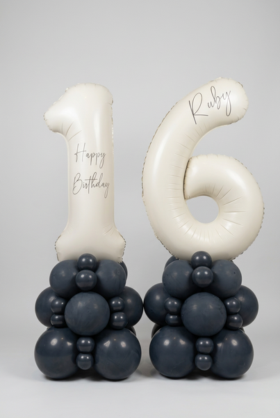 Personalised cream number 16 foil balloons on two charcoal grey balloon towers, with Happy Birthday wording and the name Ruby.