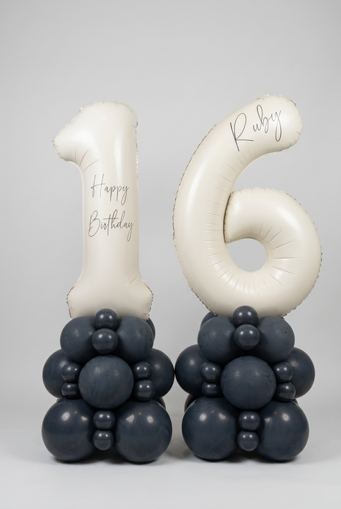 Personalised cream number 16 foil balloons on two charcoal grey balloon towers, with Happy Birthday wording and the name Ruby.