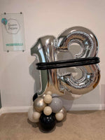 A large metallic silver foil balloon shaped like the number 3 stands on a cluster of black, white, and silver latex balloons against an indoor wall.