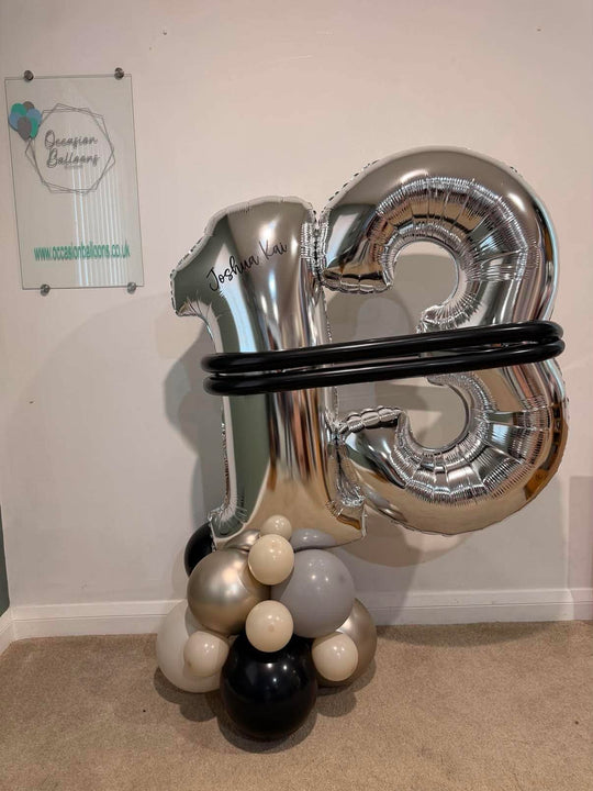 A large metallic silver foil balloon shaped like the number 3 stands on a cluster of black, white, and silver latex balloons against an indoor wall.