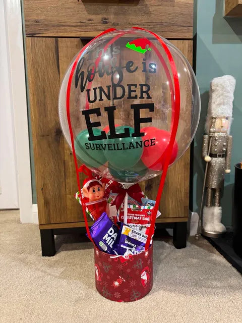 A holiday-themed gift basket with a clear balloon that reads 'Your house is UNDER ELF SURVEILLANCE' with red and green accents, a small elf figurine, and assorted treats in a red patterned base, set in a home environment.