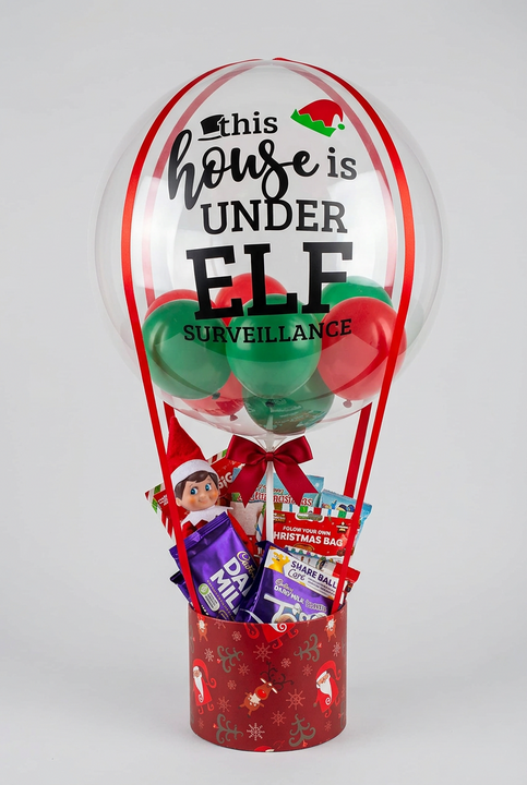 Personalised elf surveillance balloon with red and green mini balloons above a Christmas hamper with an elf, personalised bottle, treats and games.