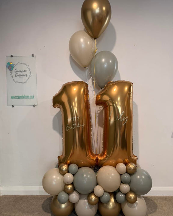 Gold number 11 balloons personalised with Lily and Happy Birthday, on a cream, grey and gold base with three matching helium balloons above.