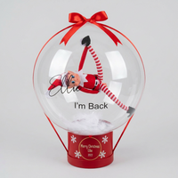 A personalized clear Christmas ornament balloon with a red ribbon bow and base, featuring a small Elf on the Shelf inside. The balloon is printed with the name 'Ellie' and the phrase 'I'm Back', and the red base is printed with 'Merry Christmas 2022'.