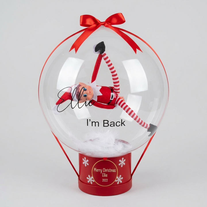 A personalized clear Christmas ornament balloon with a red ribbon bow and base, featuring a small Elf on the Shelf inside. The balloon is printed with the name 'Ellie' and the phrase 'I'm Back', and the red base is printed with 'Merry Christmas 2022'.