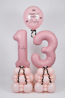Pink 13th birthday balloon decoration set featuring a large number 13 balloon, a round balloon reading 'Happy 13th Birthday Marley,' and two balloon clusters at the base tied with black ribbons, displayed indoors against a white wall.