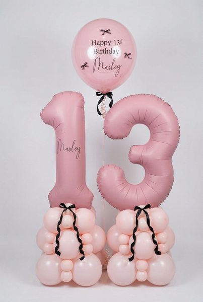 Pink 13th birthday balloon decoration set featuring a large number 13 balloon, a round balloon reading 'Happy 13th Birthday Marley,' and two balloon clusters at the base tied with black ribbons, displayed indoors against a white wall.