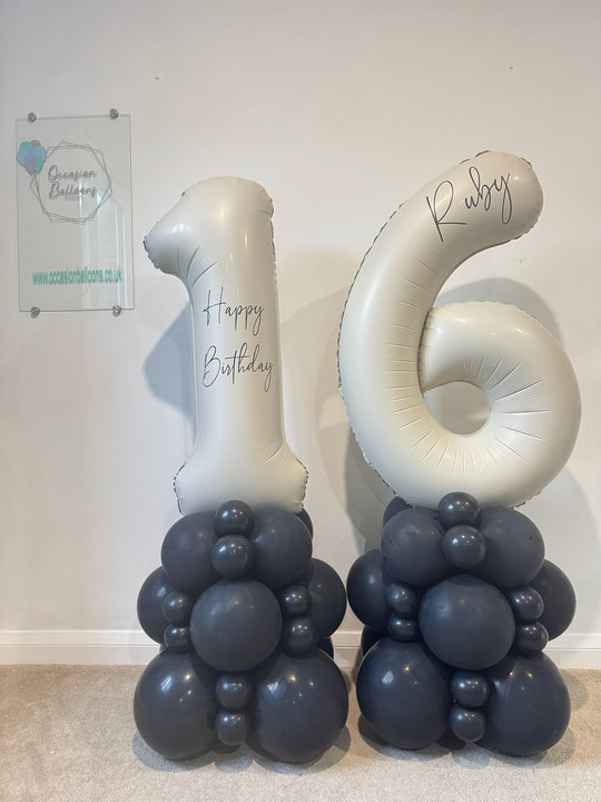 Two large metallic silver foil balloons shaped like the numbers 1 and 6, each printed with 'Happy Birthday' or 'Ruby', mounted on dark gray balloon clusters and standing against a plain indoor wall.