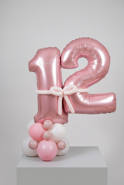 Metallic pink number 12 balloons personalised with Marley, joined by a pale pink balloon bow on a pink and white balloon base.
