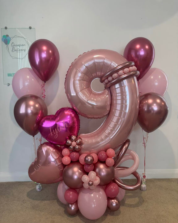 Pink number 9 balloon with personalised heart, rose gold heart, balloon flowers and a pink and berry base, beside two matching helium bouquets.