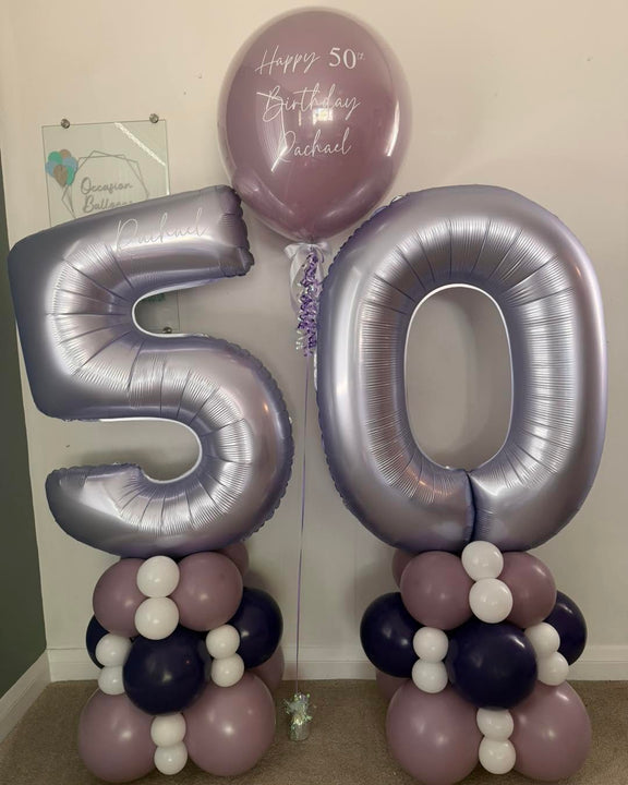 Lilac number 50 balloons on mauve, deep purple and white towers, with a personalised 24-inch birthday bubble balloon between them.