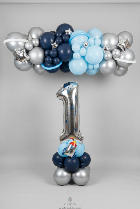 Silver number 1 balloon with a rocket on a blue and silver base, beneath a navy, light blue and silver garland with stars and balloon planet rings.